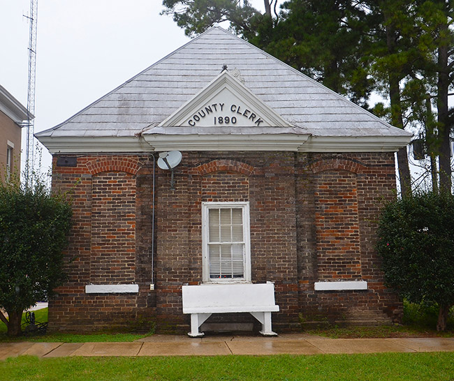 Single-story brick "County Clerk 1890" building with bench on sidewalk