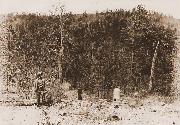 White man in hat standing next to rain gauge in wooded area