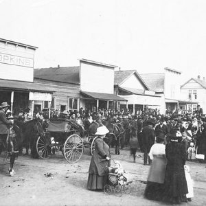 Crowded street with men women and horse drawn carriages and storefronts and one woman with a toddler in a baby carriage