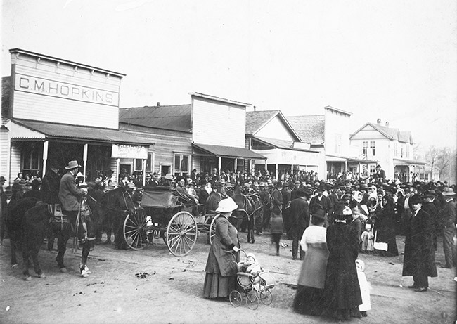 Crowded street with men women and horse drawn carriages and storefronts and one woman with a toddler in a baby carriage