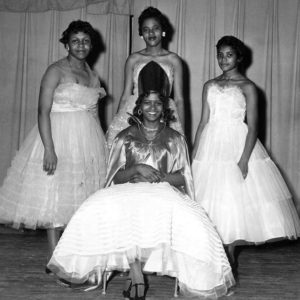 Young African-American woman sitting with crown and dress with three young African-American women in dresses behind her