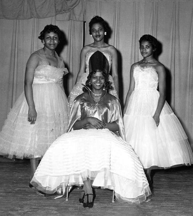 Young African-American woman sitting with crown and dress with three young African-American women in dresses behind her