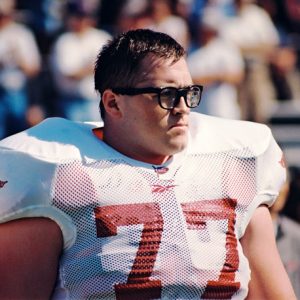 White man with glasses in white football uniform with red number 77 focusing on game in progress