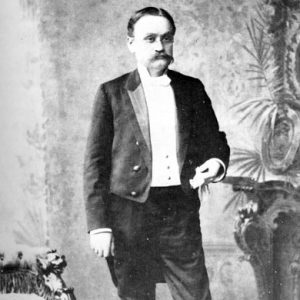 White man with mustache in tuxedo posing near fireplace and chair