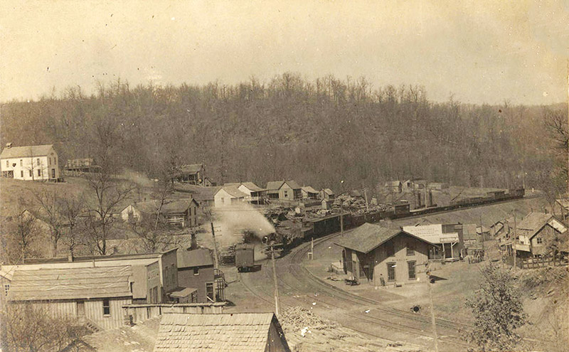 Steam locomotive passing through town on curved railroad with various houses and buildings