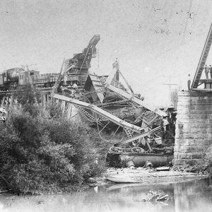 Damaged steel truss bridge with collapsed section and train in debris between near stone support