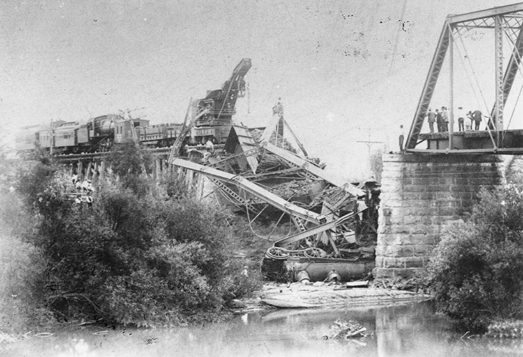 Damaged steel truss bridge with collapsed section and train in debris between near stone support