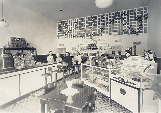 two people sitting at the counter and one person standing behind it inside a restaurant with display cases