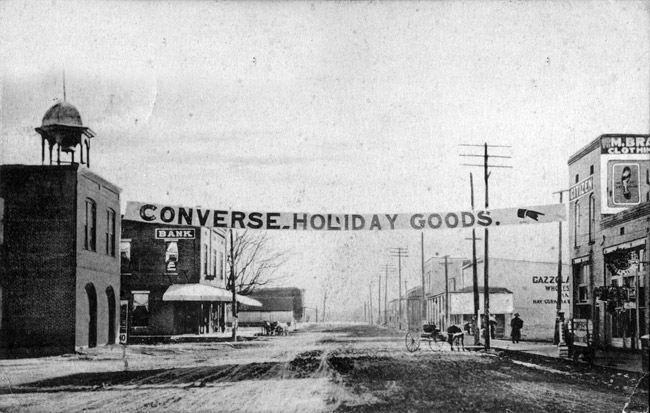 Dirt road with "Converse holiday goods" banner hanging over it between two buildings