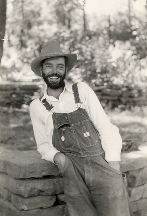 White man with beard in hat and overalls smiling while leaning on brick wall