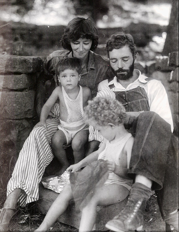 White man with beard in overalls with wife and two small children sitting next to brick wall