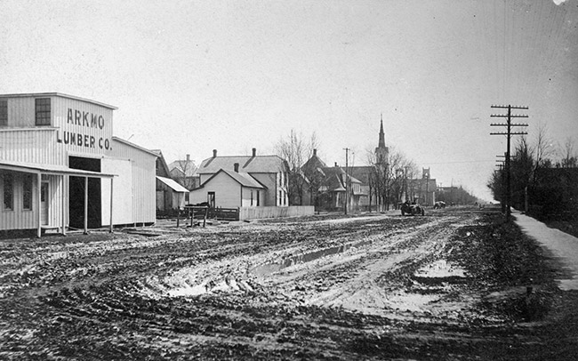 Lumber barn multistory houses and church with steeple on muddy dirt road