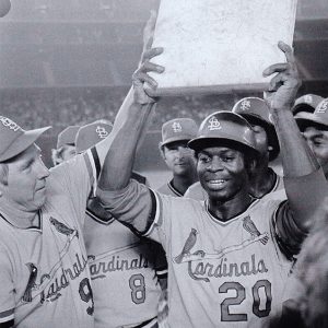 African-American man in baseball uniform holding rectangular object amid crowd of fellow players