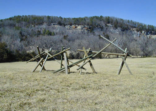 various wooden posts posed at perpendicular angles in a field with a tree covered hill in background