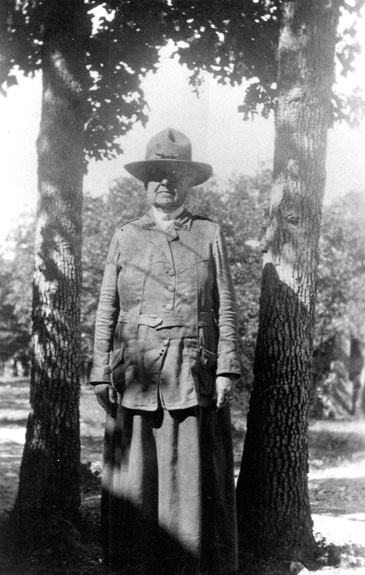 Old white woman in military-style uniform with hat standing in between two trees