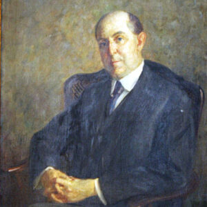 Balding white man in suit and tie sitting in a wooden chair