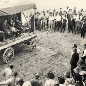 Group of white men in hats standing around wagon stage with group of white men in suits sitting on it with small table