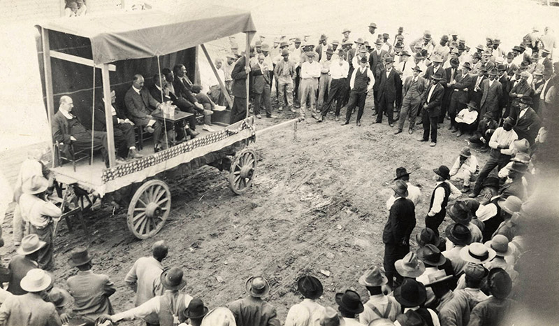 Group of white men in hats standing around wagon stage with group of white men in suits sitting on it with small table