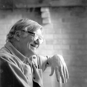 Old white man with glasses smiling with brick wall in the background