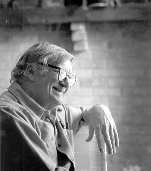 Old white man with glasses smiling with brick wall in the background