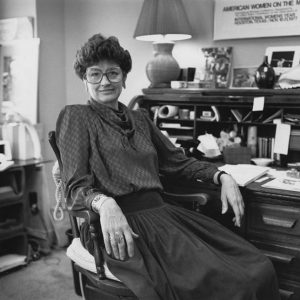 White woman with glasses wearing blouse and skirt sitting at desk