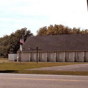 Side view of single-story church building with steeple on A-frame roof with flag poles sign on street and parking lot