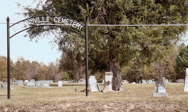 Iron arch with text in cemetery with gravestones and tree behind it