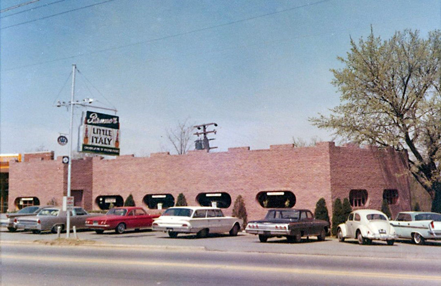 Brick building with oval shaped windows with cars in parking lot and hanging "Little Italy" sign on two lane street
