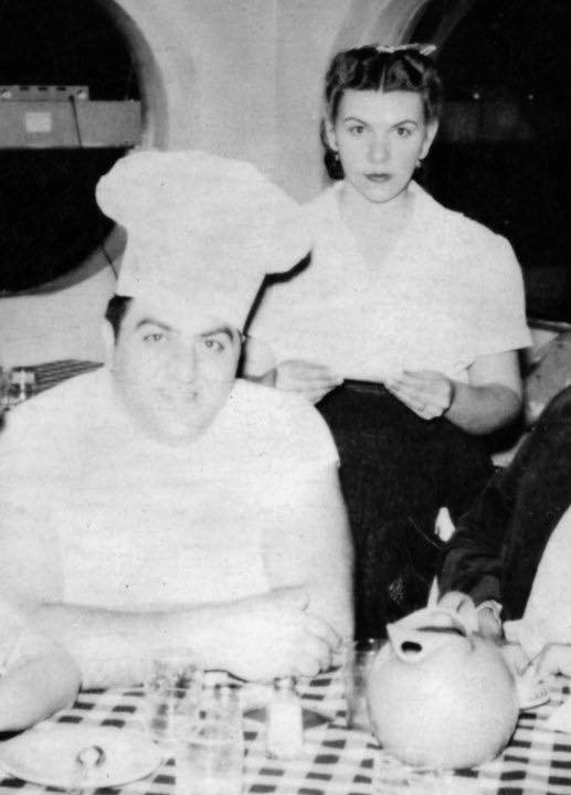 Italian-American man in chef's uniform and hat with white woman in short sleeve shirt inside restaurant with oval windows