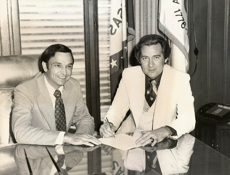 Two white men in suits sitting at desk in office with flags and telephone on a table behind them