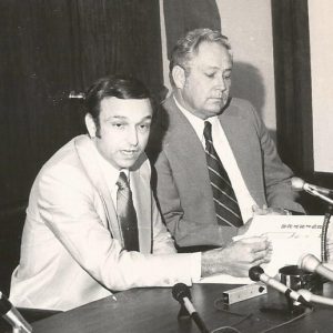 White men in suits sitting at table with microphones