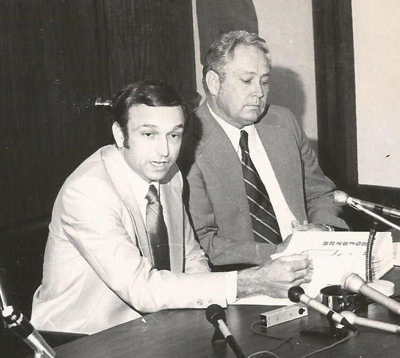 White men in suits sitting at table with microphones