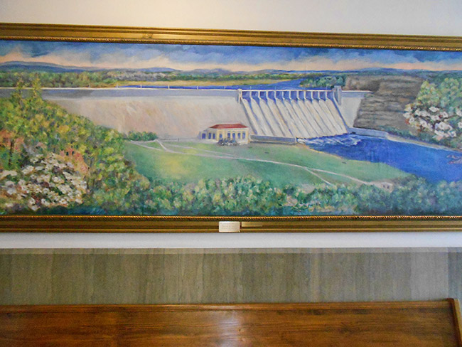 framed painting on wall showing a concrete dam with river and surrounding countryside