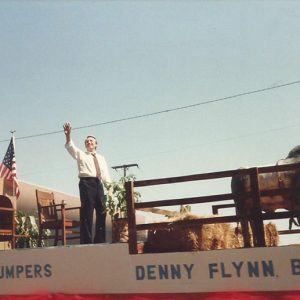 White man in shirt and tie waving to crowd from parade float with office furniture and bull on it