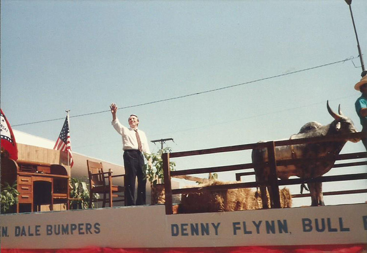 White man in shirt and tie waving to crowd from parade float with office furniture and bull on it