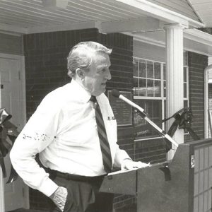 Old white man in shirt and tie speaking at lectern outside brick building with people watching behind portrait on easel to the right