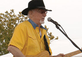 Older white man in a yellow shirt and black hat playing an electric guitar on stage in front of a microphone
