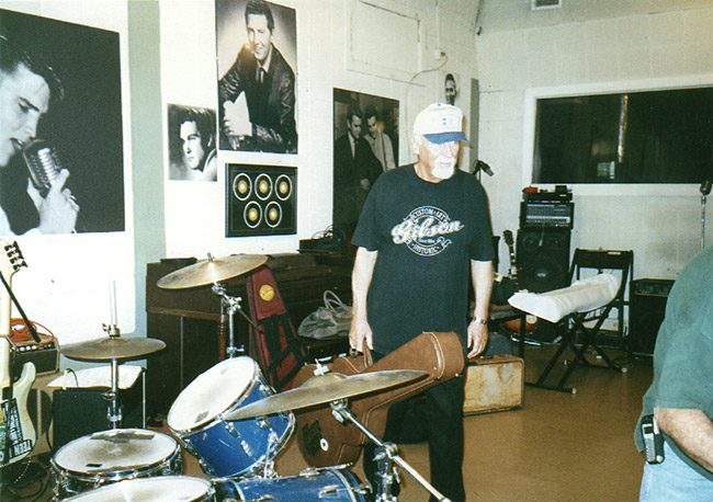 Older white man with ball cap and guitar case standing in recording studio with drum set and photographs on the walls