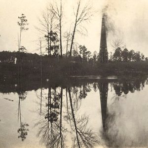 Trees and erupting oil derrick reflected in large reservoir of liquid