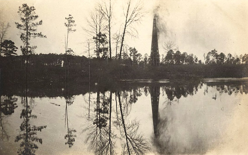 Trees and erupting oil derrick reflected in large reservoir of liquid