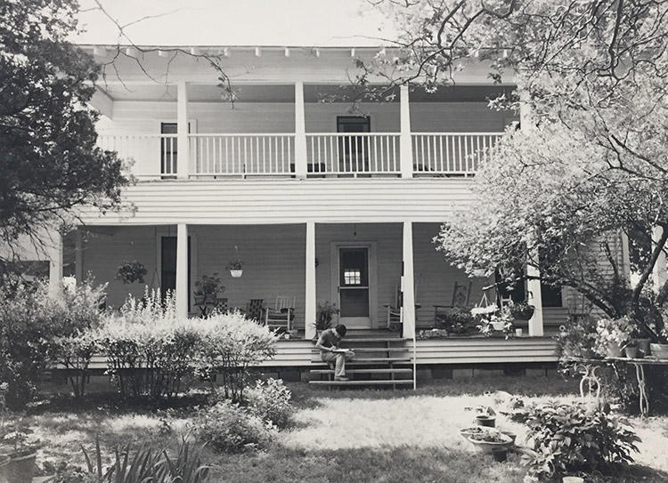 White man sitting on steps of two-story house with covered porch and balcony