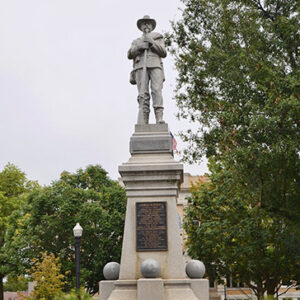 Statue of soldier holding gun on tall stone pedestal with plaques on front in park with flowers at its base