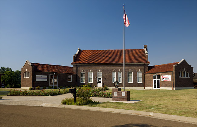 Multistory brick building with single-story wings on grass with flag pole and bell on brick pedestal next to paved driveway