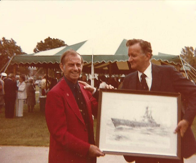 White man in red jacket handing a framed photograph of naval ship to white man in suit and bow tie with a tent and people behind them