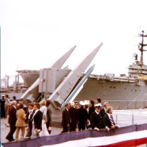 Crowd of men and women standing on dock with missile launcher and aircraft carrier in background