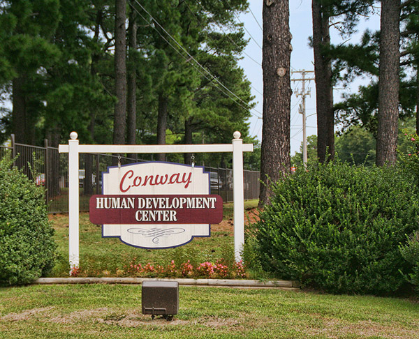white wooden post sign reading "Conway Human Development Center" amid trees with chain link fence in background