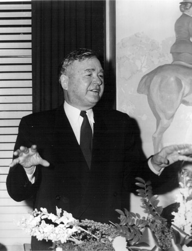 white man in suit and tie gestures while speaking  in front of painting and window obscured by curtains, blinds