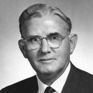 Portrait of wide-eyed white man with slight smile wavy combed hair suit jacket tie and glasses