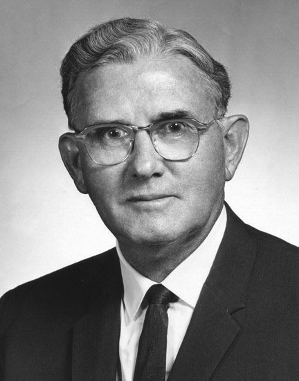 Portrait of wide-eyed white man with slight smile wavy combed hair suit jacket tie and glasses