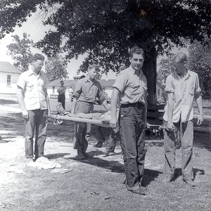 Young white men carrying white man on stretcher with white observers and building in background